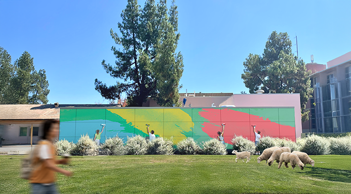 Four people use paint rollers to apply bright green, yellow, and red swaths of color to a large outdoor mural on a utility structure. Sheep graze on the lawn in the foreground, and a student walks by holding a drink. The scene is set on the UC Davis campus, with trees and buildings in the background under a clear blue sky.
