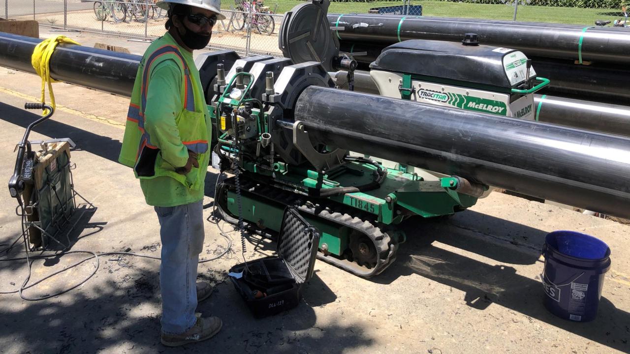 Sections of the new hot water pipe are fused together to create the length we need before being installed. Man standing by a machine fusing two sections of pipe together